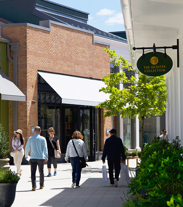 Shoppers stroll through a modern open-air outlet mall, with storefronts and a sign for The Bicester Collection.